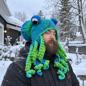May include: A crochet hat featuring a blue and green variegated yarn pattern with two large, round, white and black eyes on top and multiple long, dangling, bright green and blue curly tentacles extending from the sides of the head covering.