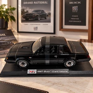 May include: A black 1987 Buick Grand National die-cast model car mounted on a black display base. The model features detailed silver wheels, a glossy finish, and a clear tape strip across the roof. The base includes a label reading Maisto 1987 Buick Grand National.