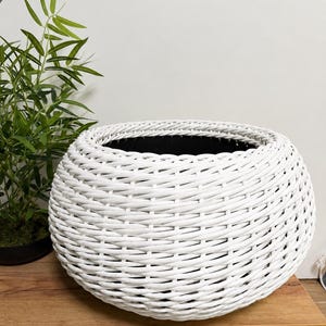 Peut inclure: Un grand panier rond blanc tissé avec un intérieur noir. Le panier est fait d'un matériau blanc épais et étroitement tissé. Le panier est posé sur une surface en bois. Une plante verte est visible en arrière-plan.