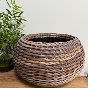 May include: A round, woven wicker basket featuring a natural brown finish with a tight, diagonal weave pattern. The basket has a wide, open top and a deep, rounded body with a dark interior lining visible inside the structure of the container.