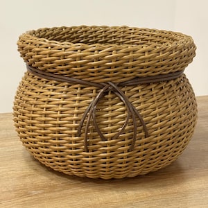 May include: A round, light brown woven basket with a thick, braided rim. The basket features a dark brown, thin cord wrapped around the upper section, tied into a decorative knot at the front, creating a textured, rounded, and open-top container.