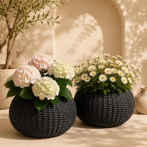 May include: Two round, dark gray woven planters with a textured finish. One planter holds a cluster of pale pink and white hydrangea flowers with green leaves. The other planter contains a dense arrangement of small white daisies with yellow centers.