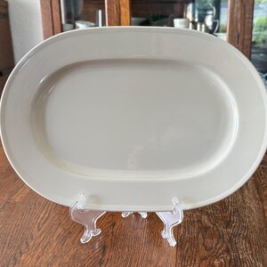 May include: A solid off-white oval serving platter with a smooth, glossy finish and a wide, flat rim. The platter is displayed upright on a clear plastic stand, showcasing its simple, clean design and uniform light cream colour throughout.