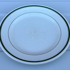 May include: A round white ceramic dinner plate featuring a thin dark green band encircling the rim. The centre of the plate shows light surface wear and minor scratches consistent with use, while the overall design remains simple and functional.