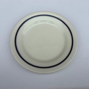 May include: A round, off-white ceramic plate featuring a smooth, glossy finish. The plate is decorated with a simple, thin, dark navy blue circular band that runs along the inner rim, providing a clean and understated contrast to the light base.