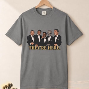 May include: A grey short-sleeve t-shirt featuring a graphic print of four men in black suits and bow ties standing alongside a grey alien. Below the figures, the text reads "THEY'RE HERE." in a bold, gold-colored serif font.