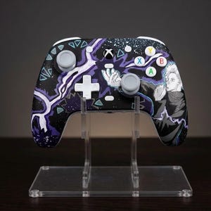 May include: A custom Xbox wireless controller featuring a black base with purple lightning patterns, white geometric shapes, and an anime-style character illustration on the right side. It includes standard gray analog sticks, a white D-pad, and buttons.
