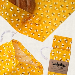 May include: A yellow beeswax food wrap set featuring a repeating bee pattern. The set includes a large wrap covering a loaf of bread, a drawstring pouch, and a folded wrap with a paper label reading UP NORTH BEESWAX FOOD WRAPS Keeps Food Fresh.