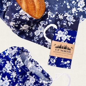 Puede incluir: Un juego de envoltorios de cera de abeja azul oscuro con un patrón floral blanco. Un envoltorio está doblado y asegurado con una banda de papel color canela etiquetada UP NORTH BEESWAX FOOD WRAPS, cubriendo parcialmente un pan redondo.