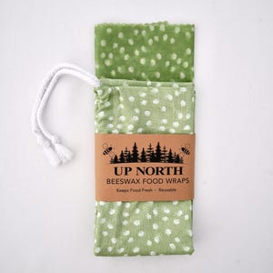 May include: A folded green fabric food wrap with a white polka dot pattern, secured by a brown paper band labeled UP NORTH BEESWAX FOOD WRAPS. The band features a black graphic of bees and pine trees, with two white braided cotton drawstrings attached.