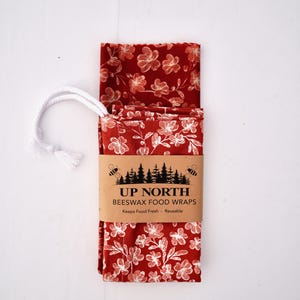 May include: A folded red beeswax food wrap featuring a white floral pattern. A tan paper band wraps around the center with the black text UP NORTH BEESWAX FOOD WRAPS, Keeps Food Fresh, Reusable, and a graphic of trees and bees. A white rope is attached.