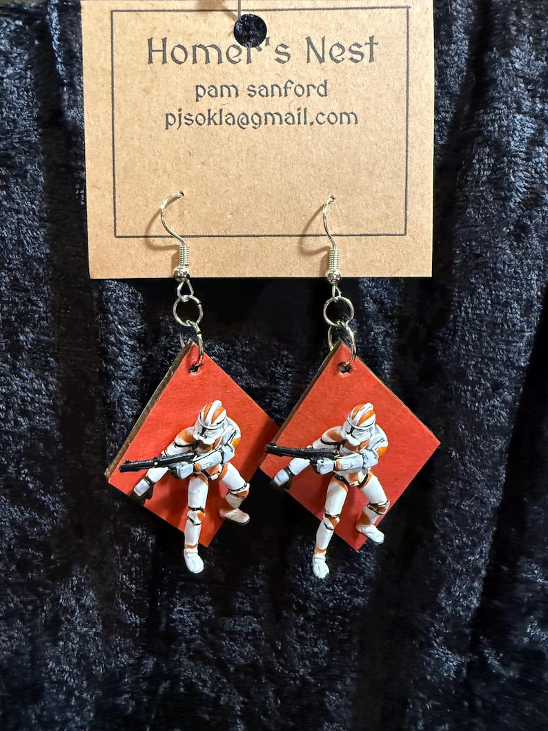 May include: A pair of handmade earrings featuring miniature figures of clone troopers on red diamond-shaped backgrounds. The figures are white and orange, holding black rifles. The earrings hang from silver hooks.