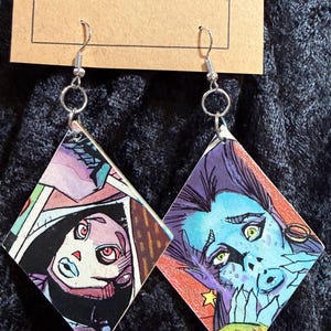 May include: A pair of diamond-shaped earrings with illustrations of stylized faces. The earrings feature silver-colored hooks and rings. One earring has a face with red eyes and black hair, the other has a blue face with green eyes.