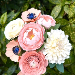 May include: A bouquet of paper flowers in shades of pink, peach, white, and yellow. The arrangement includes peonies, anemones, and a dahlia, with green paper leaves. The flowers are set against a backdrop of green foliage.
