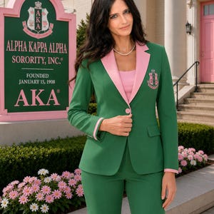 Pode incluir: Um blazer verde com lapelas e punhos cor-de-rosa, usado sobre um top cor-de-rosa. O blazer apresenta um fecho de um botão e um emblema bordado da Alpha Kappa Alpha Sorority, Inc. no peito. O conjunto inclui calças verdes a combinar.