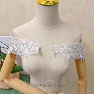 May include: Two detachable off-the-shoulder sleeves made of sheer white tulle fabric featuring intricate white floral embroidery and scalloped edges. The pieces are displayed on a mannequin torso with the text LuneBridalAccents visible.