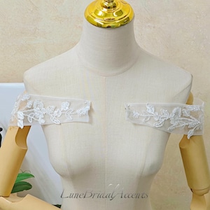 May include: A pair of off-the-shoulder bridal sleeves made of sheer white tulle fabric featuring intricate white floral embroidery with delicate beadwork. The sleeves are displayed on a beige mannequin with a gold-toned neck cap.