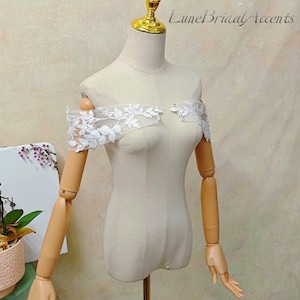 May include: A beige mannequin torso featuring off-the-shoulder white lace sleeves with a detailed floral and leaf embroidery pattern. The lace is sheer and delicate, extending across the upper arms. The text LuneBridalAccents is visible.