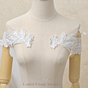 May include: A pair of white off-the-shoulder lace appliques featuring intricate floral and leaf embroidery on sheer mesh fabric. Each piece is designed to be attached to a garment, showcasing detailed stitching and delicate scalloped edges.