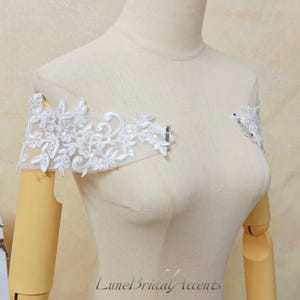 May include: A pair of white off-the-shoulder lace appliques featuring intricate floral embroidery on sheer mesh fabric. The delicate white lace pieces are positioned on a beige mannequin, showcasing detailed leaf and petal patterns.