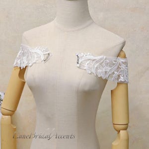 May include: A pair of detachable off-the-shoulder sleeves featuring white floral lace embroidery on sheer tulle fabric. Each sleeve is finished with a silver-toned metal pin attachment for securing to a garment. The text LuneBridalAccents is visible.