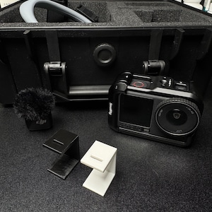 May include: A black action camera in a protective cage, a small fuzzy microphone, and two L-shaped plastic mounts in black and white. The camera features a circular lens and a screen with the text ACTION. A black hard-shell equipment case is behind.