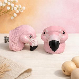 Könnte beinhalten: Zwei rosa Schrankknöpfe in Form eines Flamingos. Die Knöpfe haben schwarze Schnäbel und Augen, mit einer Schraube auf der Rückseite eines Knopfes. Die Flamingoköpfe blicken in verschiedene Richtungen. Die Knöpfe sind aus einem festen Material.