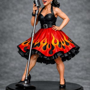 Op de afbeelding: Figuur van een vrouw in een zwarte haltertop en een rode rok met vlammen, die een microfoon vasthoudt. Ze draagt een zonnebril. De figuur staat op een zwarte basis, met een retro rockabilly thema. De rok heeft een zwarte volant.
