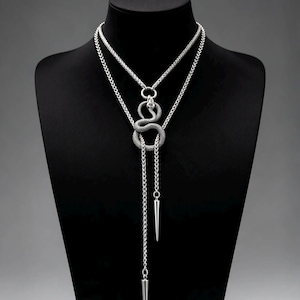 May include: A silver-toned lariat necklace featuring a textured snake pendant coiled around a circular ring. The necklace has two long, dangling chains, each finished with a smooth, pointed metal spike charm at the end, creating a layered look.