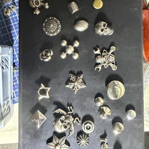K&ouml;nnte beinhalten: Eine Sammlung verschiedener silberfarbener Metallkn&ouml;pfe und Nieten in unterschiedlichen Formen, darunter Totenk&ouml;pfe, Sterne, Rosen und geometrische Muster. Diese dekorativen Verschl&uuml;sse sind auf einer dunklen Oberfl&auml;che angeordnet und zeigen vielf&auml;ltige Texturen.