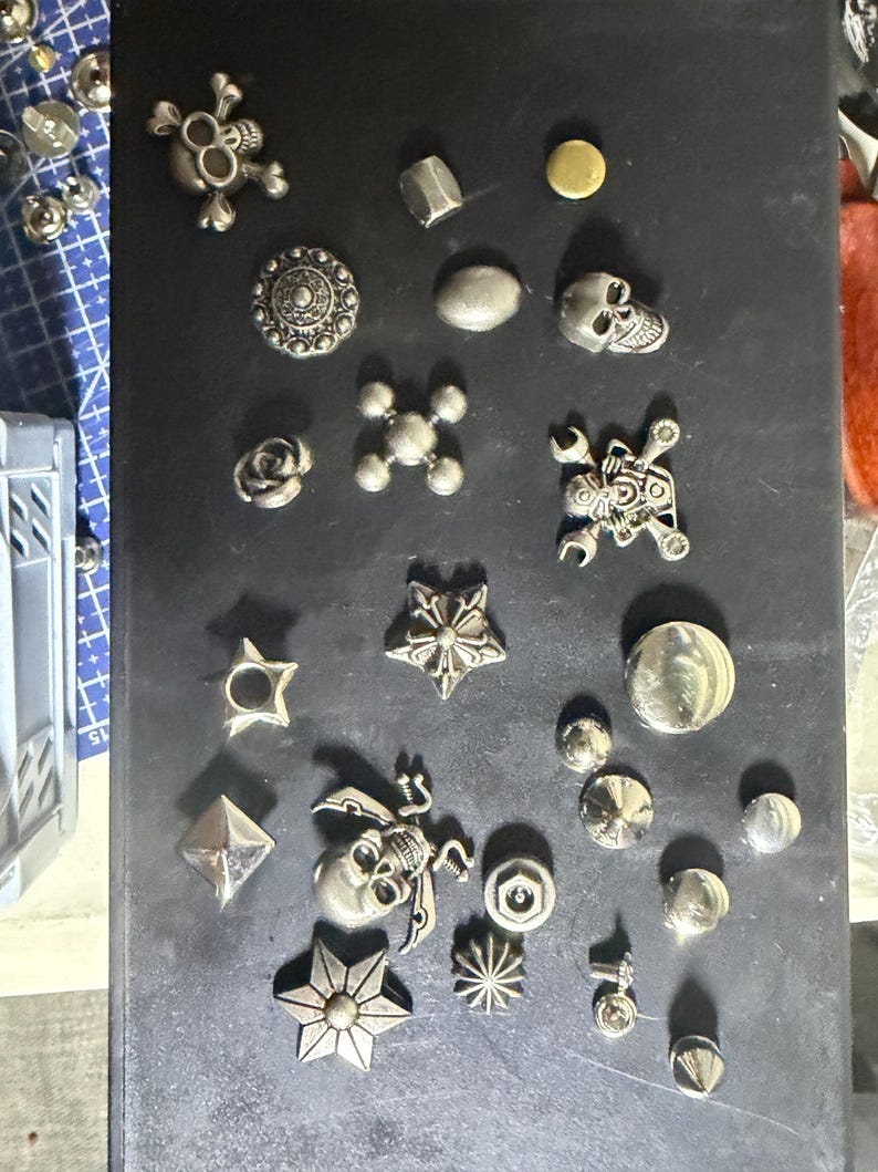 K&ouml;nnte beinhalten: Eine Sammlung verschiedener silberfarbener Metallkn&ouml;pfe und Nieten in unterschiedlichen Formen, darunter Totenk&ouml;pfe, Sterne, Rosen und geometrische Muster. Diese dekorativen Verschl&uuml;sse sind auf einer dunklen Oberfl&auml;che angeordnet und zeigen vielf&auml;ltige Texturen.