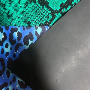 K&ouml;nnte beinhalten: Drei &uuml;berlappende Materialb&ouml;gen: eine obere Schicht mit einem leuchtend gr&uuml;nen und schwarzen Schlangenledermuster, eine mittlere Schicht mit einem leuchtend blauen und schwarzen Leopardenmuster und eine untere Schicht aus glattem, mattem schwarzem Material.