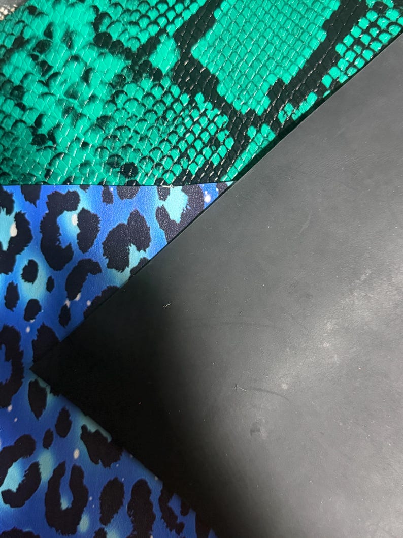 K&ouml;nnte beinhalten: Drei &uuml;berlappende Materialb&ouml;gen: eine obere Schicht mit einem leuchtend gr&uuml;nen und schwarzen Schlangenledermuster, eine mittlere Schicht mit einem leuchtend blauen und schwarzen Leopardenmuster und eine untere Schicht aus glattem, mattem schwarzem Material.