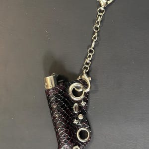 K&ouml;nnte beinhalten: Eine Feuerzeugh&uuml;lle mit dunkelviolettem und schwarzem Schlangenledermuster und silberfarbenen Metallbeschl&auml;gen, einschlie&szlig;lich einer Kette mit herzf&ouml;rmigem Verschluss, runden &Ouml;sen und kleinen dekorativen Metallnieten auf der strukturierten Oberfl&auml;che.