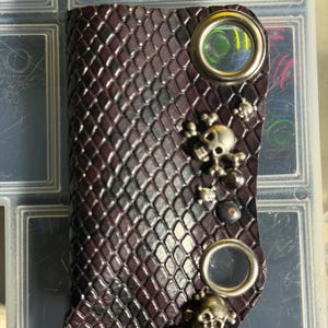 K&ouml;nnte beinhalten: Ein dunkelvioletter, strukturierter Leder-Schl&uuml;sselanh&auml;nger mit einem sich wiederholenden Rautenmuster. Das Accessoire ist mit zwei gro&szlig;en silberfarbenen Metall&ouml;sen, mehreren kleinen Totenkopf-Anh&auml;ngern und einer sechseckigen Metallmutter verziert.