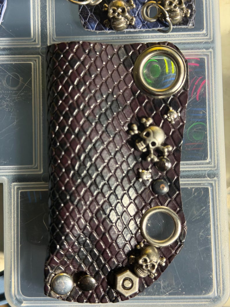 K&ouml;nnte beinhalten: Ein dunkelvioletter, strukturierter Leder-Schl&uuml;sselanh&auml;nger mit einem sich wiederholenden Rautenmuster. Das Accessoire ist mit zwei gro&szlig;en silberfarbenen Metall&ouml;sen, mehreren kleinen Totenkopf-Anh&auml;ngern und einer sechseckigen Metallmutter verziert.