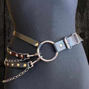 Op de afbeelding: Een zwarte leren riem met een grote zilverkleurige metalen ring in het midden. De riem is voorzien van een zilveren gesp, twee extra leren banden die met zilveren karabijnhaken aan de ring zijn bevestigd, zilveren studs en een hangende ketting.