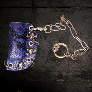 May include: A blue and black snakeskin-textured pouch featuring three silver-toned skull and crossbones charms attached to metal rings. A silver-toned metal chain with a circular ring and an additional skull charm is connected to the pouch.