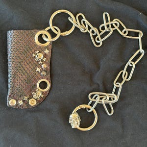 K&ouml;nnte beinhalten: Ein braunes Portemonnaie-Accessoire mit Schlangenleder-Textur, verziert mit silberfarbenen Totenkopf-Nieten, runden &Ouml;sen und einem sechseckigen Mutter-Akzent. Eine lange, schwere silberfarbene Metallkette ist befestigt und endet in einem Ring.
