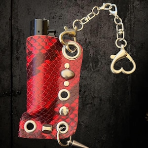 May include: A black lighter encased in a red and black snakeskin-patterned leather sleeve. The sleeve features silver-toned metal grommets, studs, and a dangling silver chain with a heart-shaped lobster clasp and a small metal spike charm.