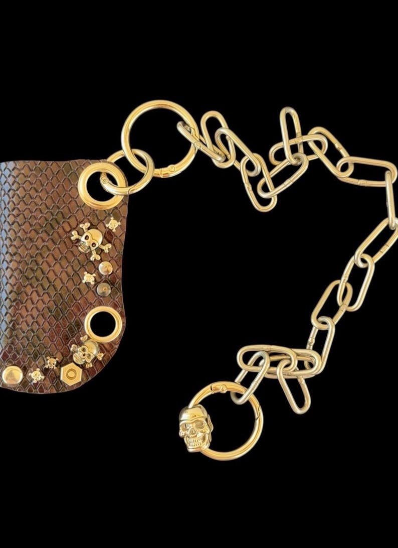 K&ouml;nnte beinhalten: Ein braunes Accessoire mit Schlangenlederstruktur, verziert mit goldfarbenen Totenkopf- und Knochennieten, einem Sechskantmuttern-Akzent und einer langen goldfarbenen Gliederkette, die in einem Ring mit kleinem Totenkopf-Anh&auml;nger endet.