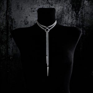 Könnte beinhalten: Eine silberfarbene Lariat-Halskette mit einem zweilagigen Ketten-Choker, der an einem zentralen Ring befestigt ist. Zwei lange, herabhängende Kettenstränge verlaufen vom Ring nach unten, jeweils mit einem spitzen, konischen Metallanhänger.