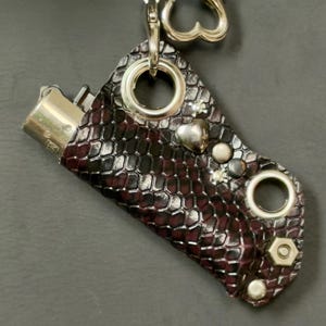 K&ouml;nnte beinhalten: Eine Feuerzeugh&uuml;lle mit einem dunkelburgunderfarbenen und schwarzen Schlangenledermuster. Die H&uuml;lle ist mit silberfarbenen Metallbeschl&auml;gen verziert, darunter zwei gro&szlig;e &Ouml;sen, ein herzf&ouml;rmiger Anh&auml;nger, kleine Nieten und eine Sechskantmutter. Eine silberne Kette mit einem herzf&ouml;rmigen Verschluss ist an der Oberseite befestigt.