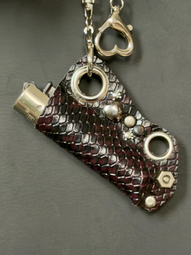 K&ouml;nnte beinhalten: Eine Feuerzeugh&uuml;lle mit einem dunkelburgunderfarbenen und schwarzen Schlangenledermuster. Die H&uuml;lle ist mit silberfarbenen Metallbeschl&auml;gen verziert, darunter zwei gro&szlig;e &Ouml;sen, ein herzf&ouml;rmiger Anh&auml;nger, kleine Nieten und eine Sechskantmutter. Eine silberne Kette mit einem herzf&ouml;rmigen Verschluss ist an der Oberseite befestigt.