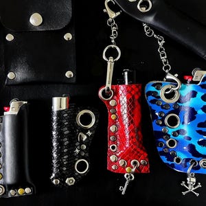 K&ouml;nnte beinhalten: Vier Feuerzeugh&uuml;llen aus Leder und Kunstleder in Schwarz, Rot und Blau-Camouflage. Jede H&uuml;lle ist mit Metall&ouml;sen, Nieten und befestigten Ketten mit Karabinerverschl&uuml;ssen versehen. Eine H&uuml;lle enth&auml;lt einen kleinen Totenkopf-Anh&auml;nger.