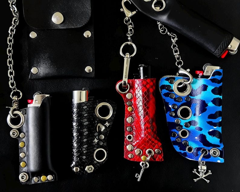 K&ouml;nnte beinhalten: Vier Feuerzeugh&uuml;llen aus Leder und Kunstleder in Schwarz, Rot und Blau-Camouflage. Jede H&uuml;lle ist mit Metall&ouml;sen, Nieten und befestigten Ketten mit Karabinerverschl&uuml;ssen versehen. Eine H&uuml;lle enth&auml;lt einen kleinen Totenkopf-Anh&auml;nger.