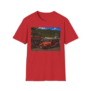 May include: Red t-shirt featuring a vibrant image of an orange Jeep parked near a roadside stand with the text "UTUADO" visible. The scene includes a mountain backdrop and colorful flags, creating a scenic view.