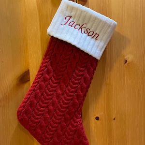 May include: Red knitted Christmas stocking with a white cuff and the name "Jackson" embroidered in red.