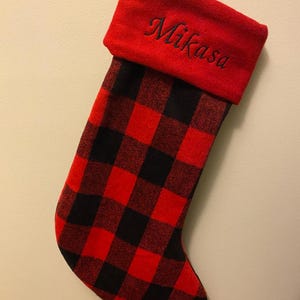 May include: Red and black buffalo plaid Christmas stocking with a red cuff. The name "Mikasa" is embroidered in black script on the cuff. The stocking has a loop for hanging.