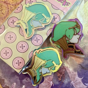 May include: Three acrylic pins featuring a stylised profile of a character with mint green hair and gold-coloured armour accents. The pins have a glossy finish and are displayed on a light-coloured backing card decorated with a pattern of pink buttons.